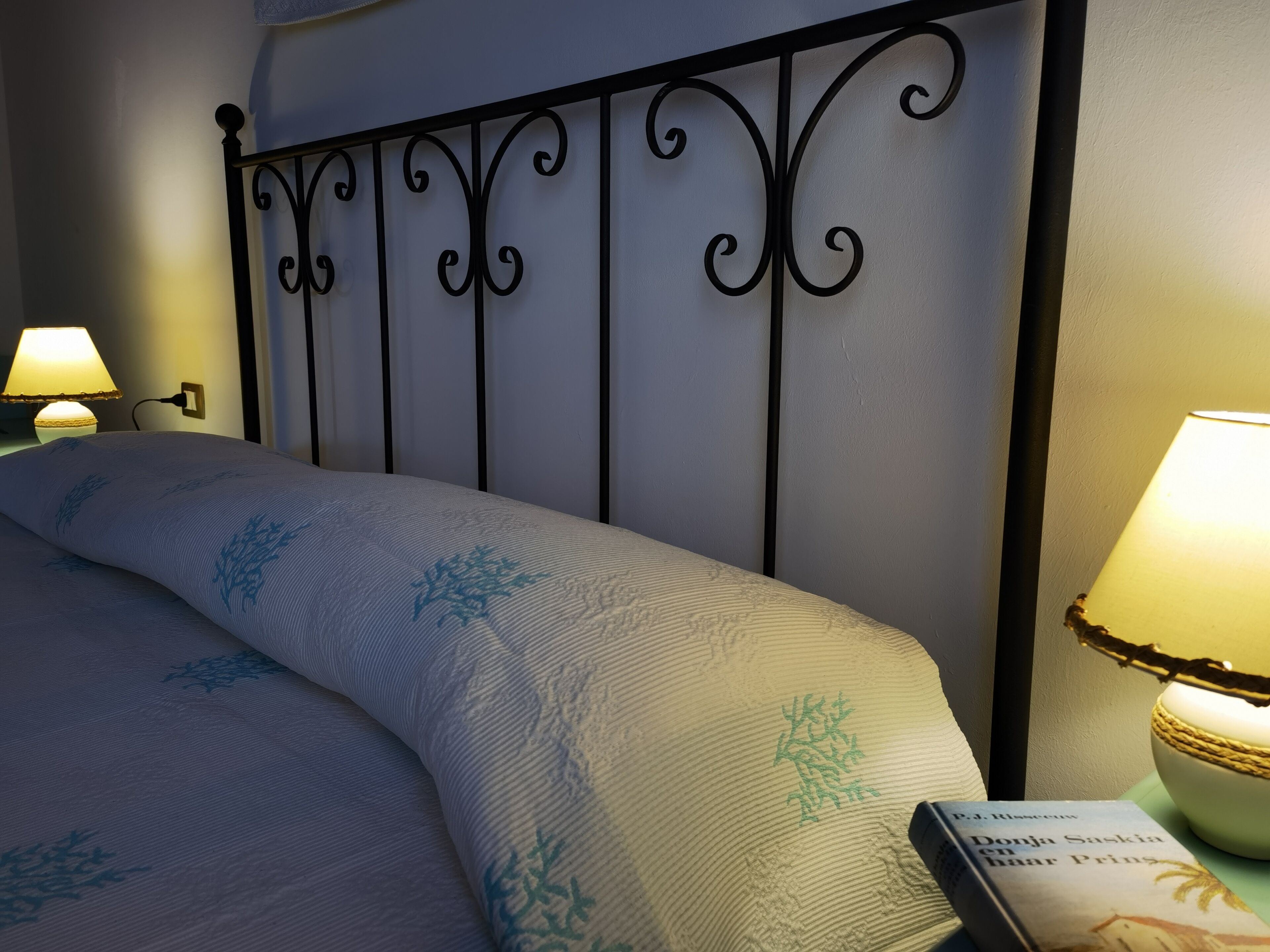 Una cama bien hecha con una colcha estampada, cabecero de metal y una lámpara de noche.