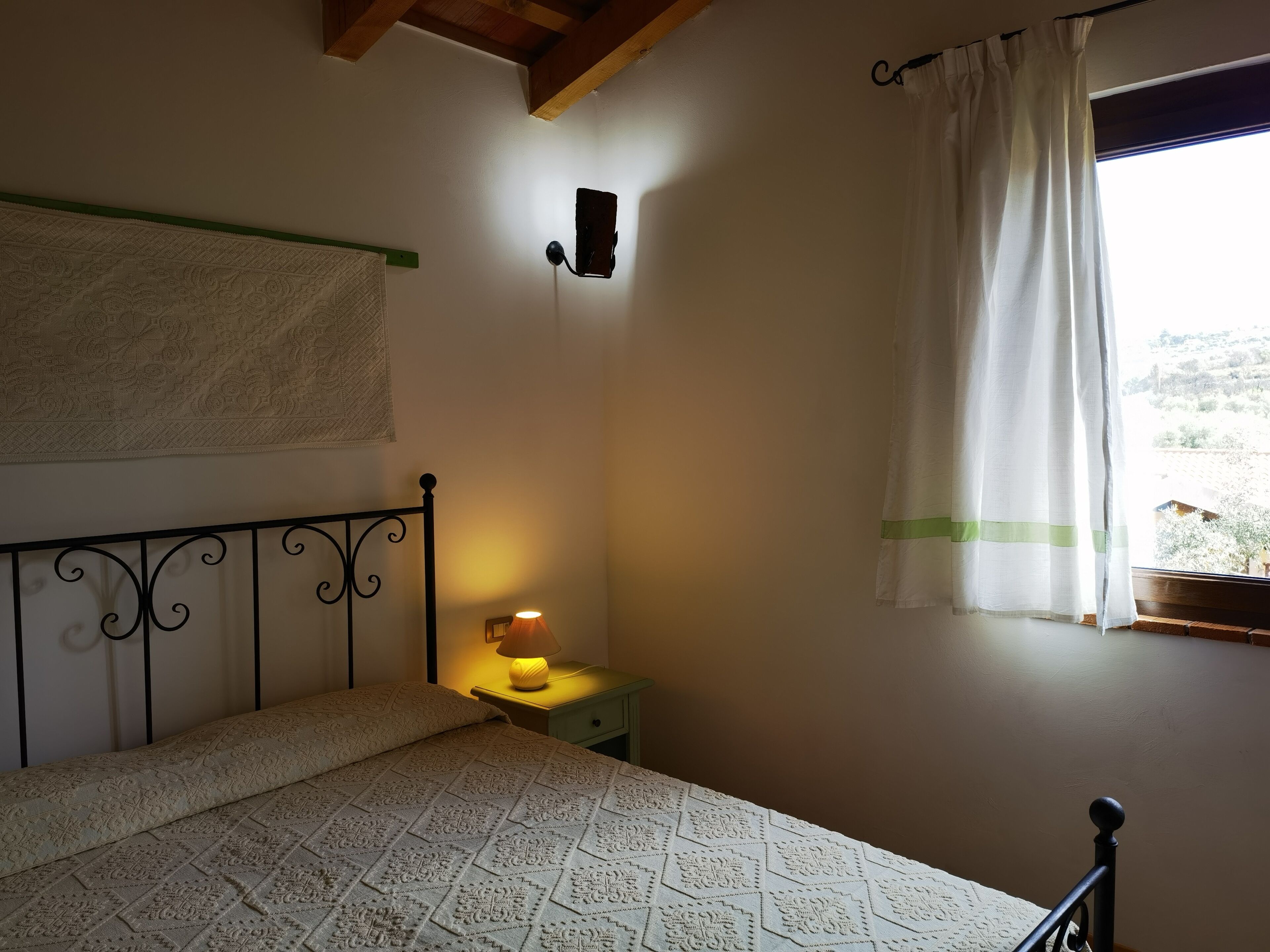 Un dormitori amb un llit, una làmpada de nit, una làmpada de paret, una finestra amb cortina i vistes a l'exterior.