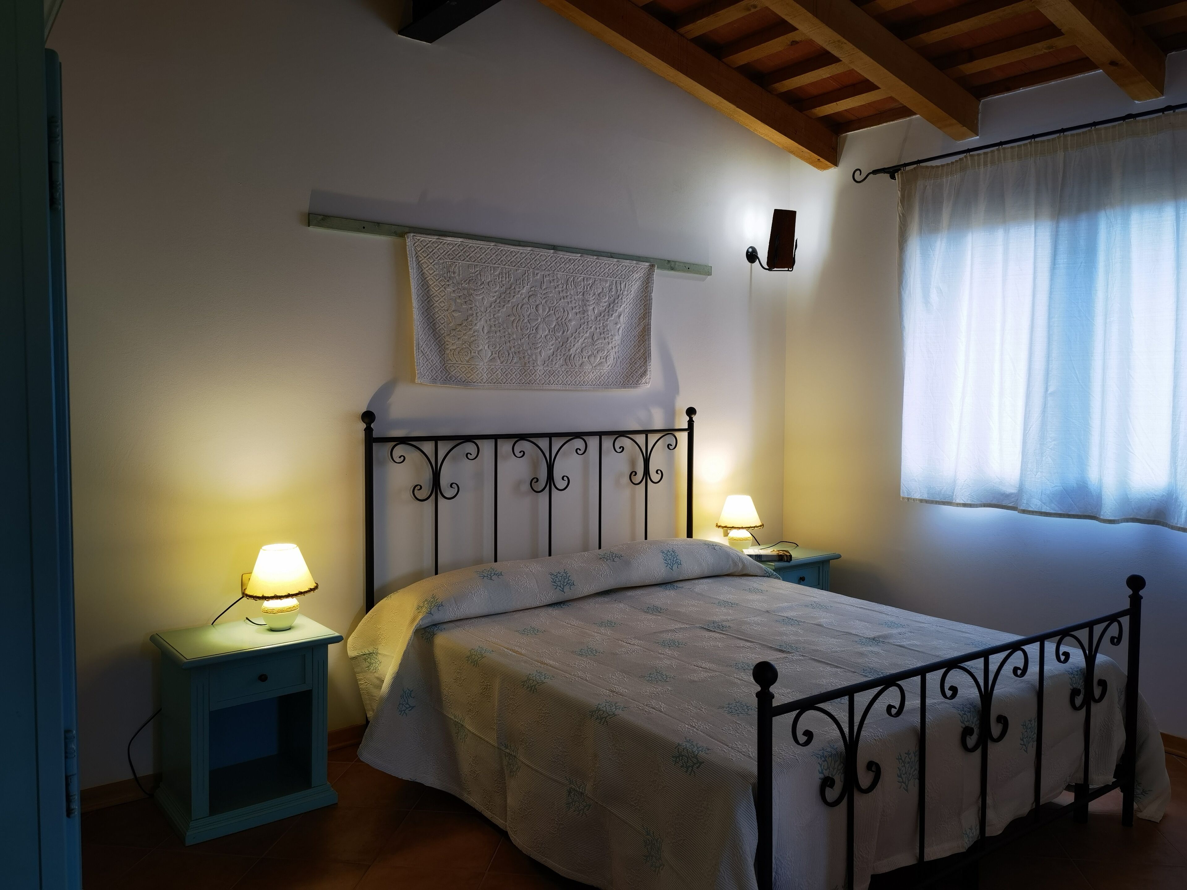 A bedroom with a bed, two bedside tables with lamps, a hanging tapestry, and a window with curtains.