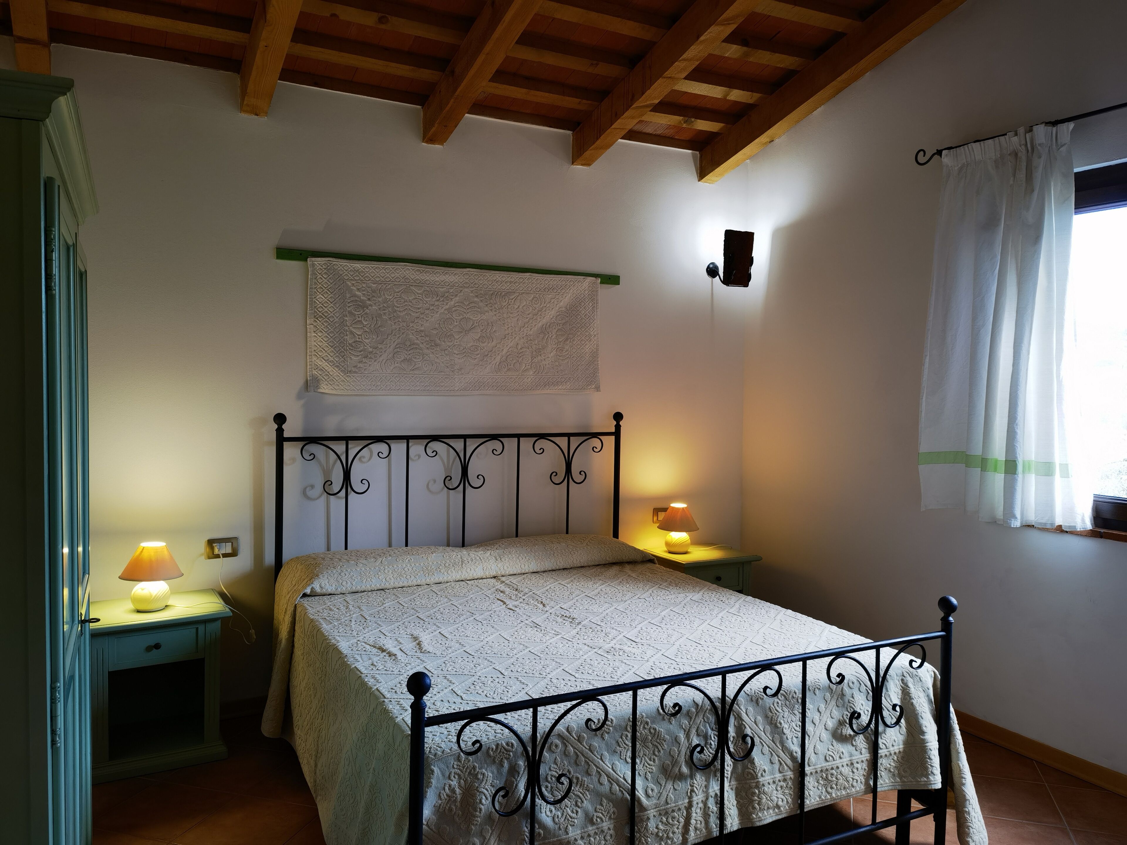 A bedroom with a bed, two bedside tables, a wall-mounted shelf, and a window with curtains.