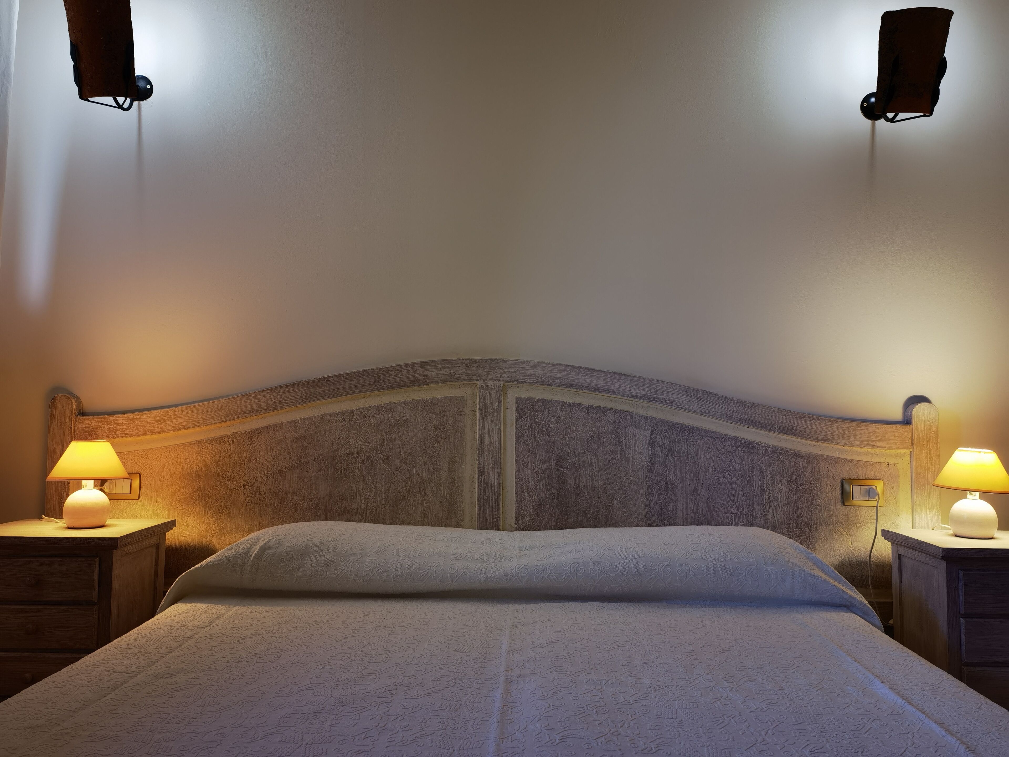 Ein ordentlich bezogenes Bett mit einem gemusterten Überwurf, zwei Nachttischlampen und einem hölzernen Kopfteil.