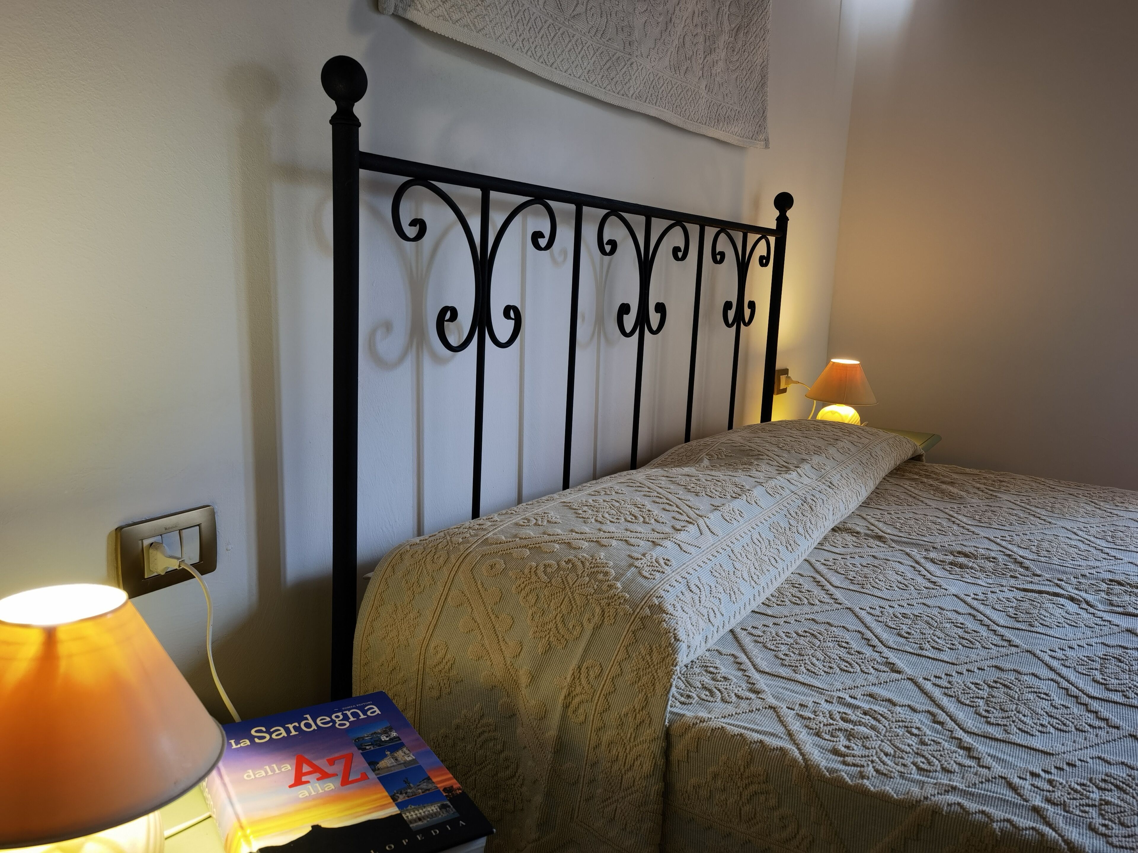 Ein ordentlich bezogenes Bett mit einem gemusterten Überwurf, ein Buch mit dem Titel 'La Sardegna dall'AZ' auf dem Nachttisch und eine Wandlampe.