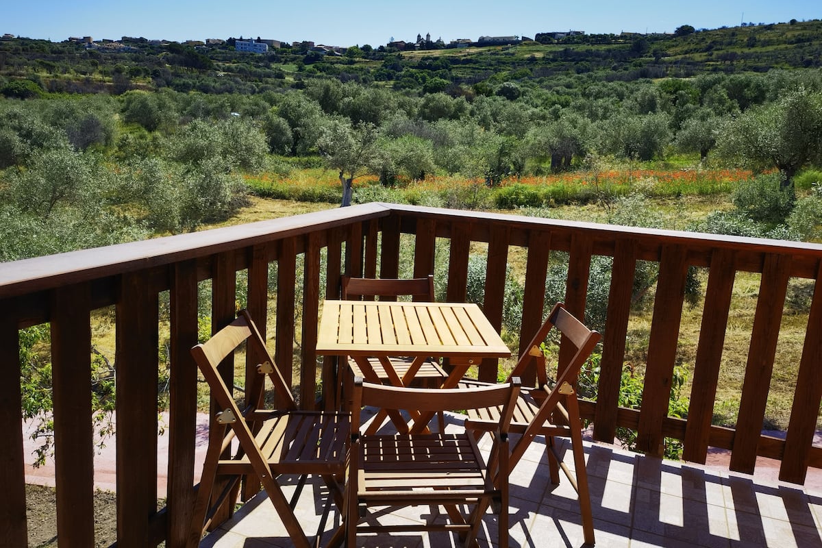 Terraza de madera con una mesa y sillas, con vistas a un paisaje ondulado con vegetación y edificios a lo lejos.