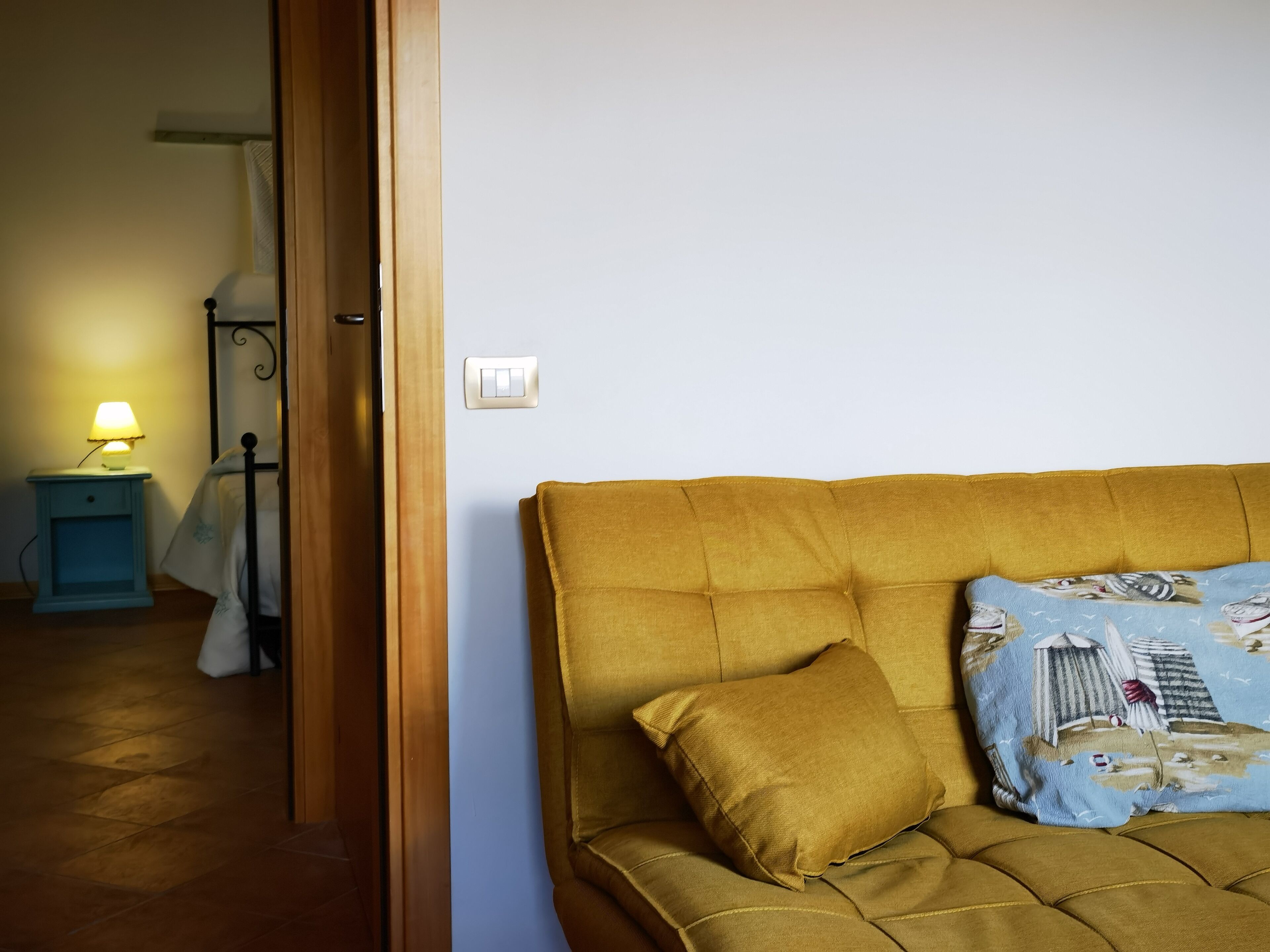 A room with a mustard-colored sofa, a patterned throw pillow, a small bedside table with a lamp, and a door leading to another room.