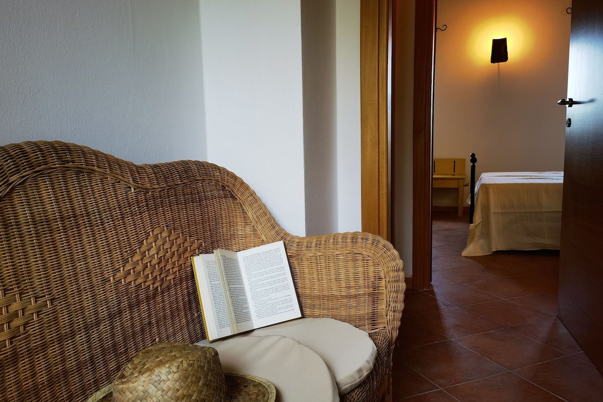 A wicker sofa with a book and a hat, a bed with a white bedspread, and a wall-mounted light fixture.