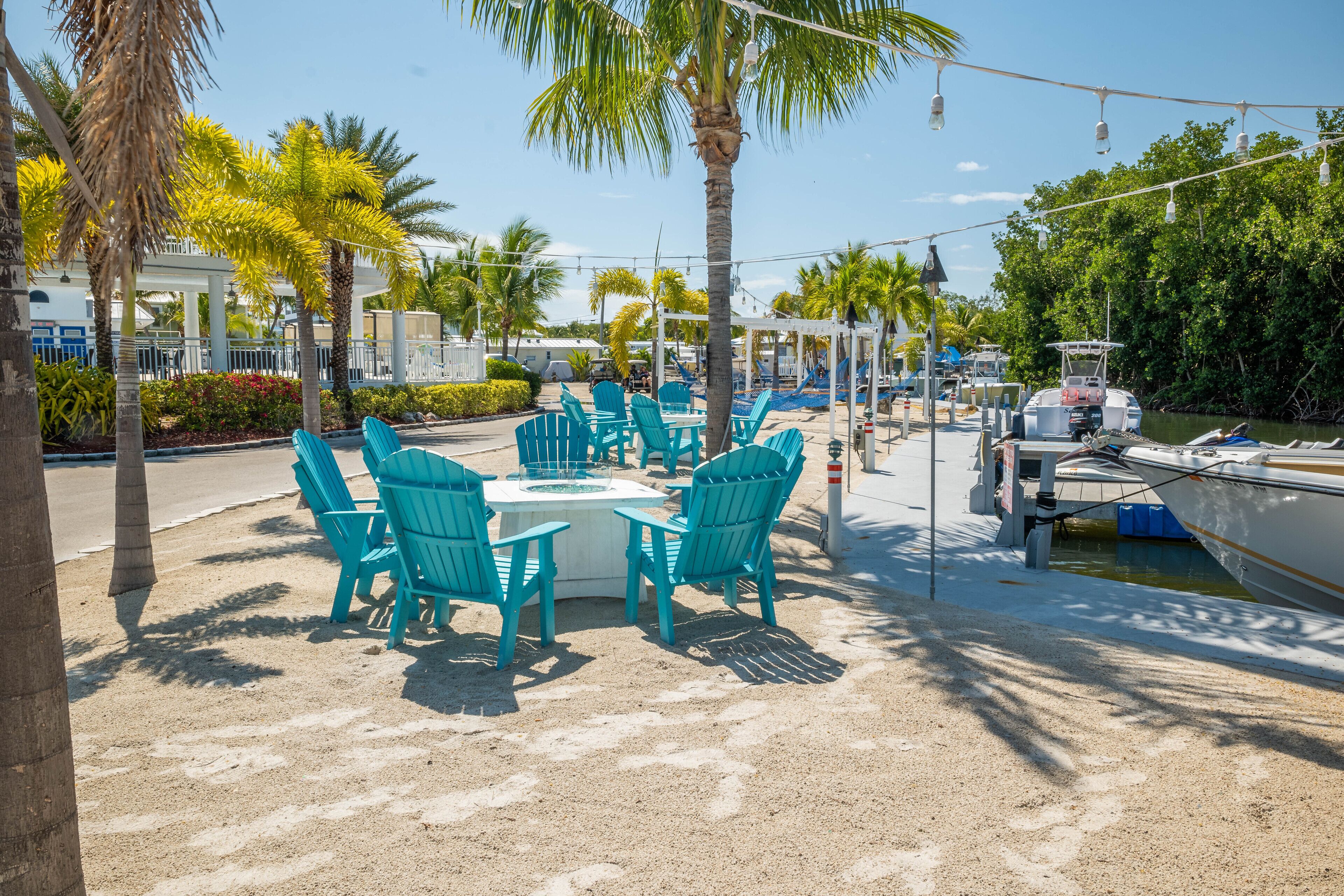 A beachfront area with turquoise chairs and tables, a boat docked nearby, and palm trees.
