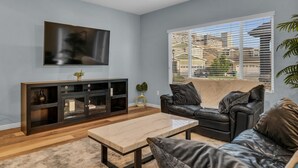 Un salon doté d’un téléviseur à écran plat, d’un meuble de rangement en bois foncé, d’un canapé en cuir, d’une table basse beige et d’une grande fenêtre avec des stores.