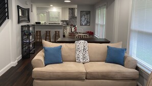 A living room with a beige sofa, two blue pillows, and a patterned throw. A kitchen with white cabinets and a dining area with a table and chairs is visible in the background. - Home Near Main Street-Covered Patio-Hot Tub! (Saint Charles)