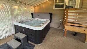 A hot tub with a wooden bench and a small step stool, surrounded by a lattice fence and string lights. - Home Near Main Street-Covered Patio-Hot Tub! (Saint Charles)