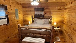 Un dormitorio rústico de cabaña de troncos con cama de madera, un banco, un ventilador de techo y una mesita de noche con lámpara.