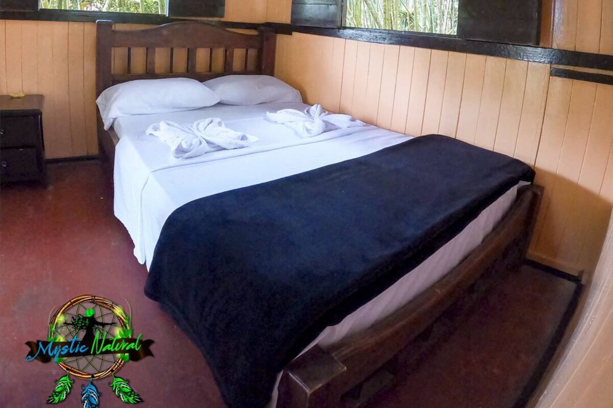Una cama individual con sábanas blancas y una colcha negra en una habitación con paredes de madera y una ventana con cortina rosa.