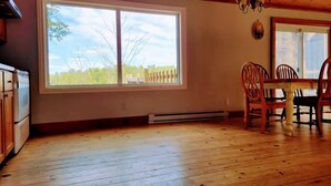 A room with wooden flooring, a dining table with chairs, a large window with a view of trees and a fence, and a ceiling fan.