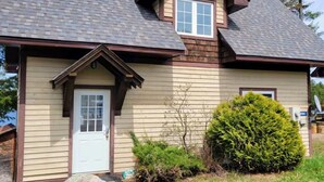 A two-story house with a gabled roof, a front door, and a small front yard with a rock and a bush.