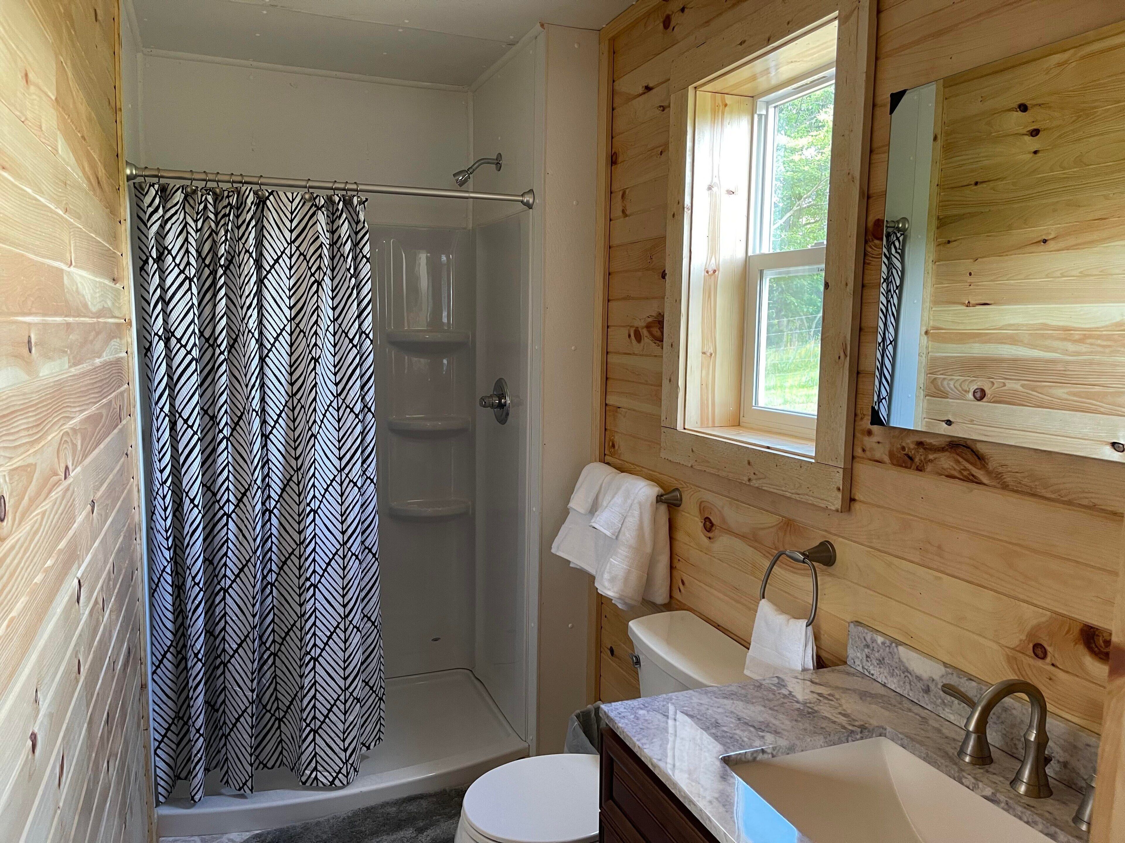 Une salle de bain rustique, aménagée dans une cabane en bois, comprenant une douche, des toilettes, un lavabo et une fenêtre laissant entrer la lumière naturelle.