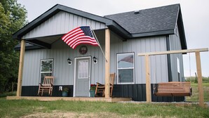 A house with a porch, a flag, and two rocking chairs. - Little House on Dairy- Near Ark- Feed Grain to Calves and Goats (Dry Ridge)