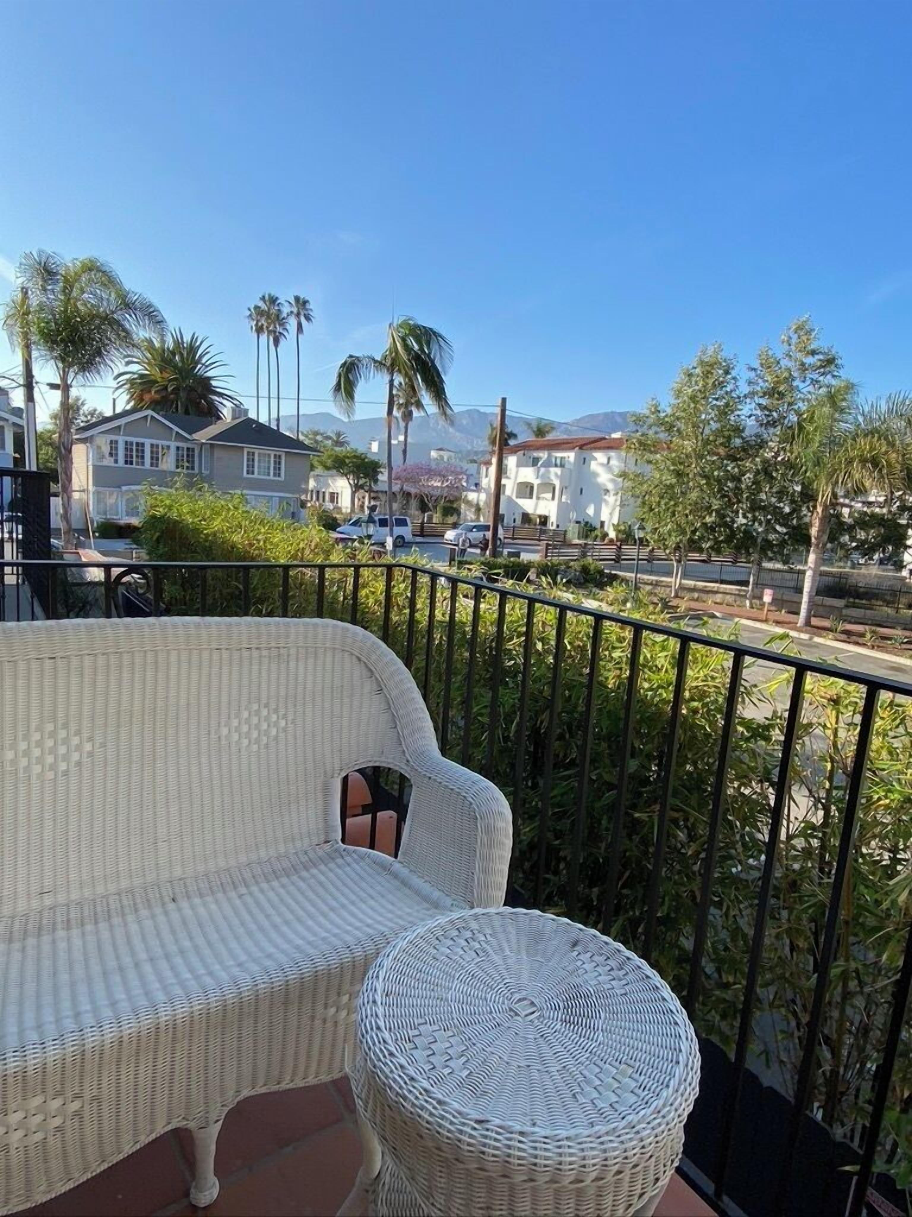 Ein Balkon mit Rattan-Sofa und Hocker, einem Metallgeländer und Blick auf Wohnhäuser und Palmen.