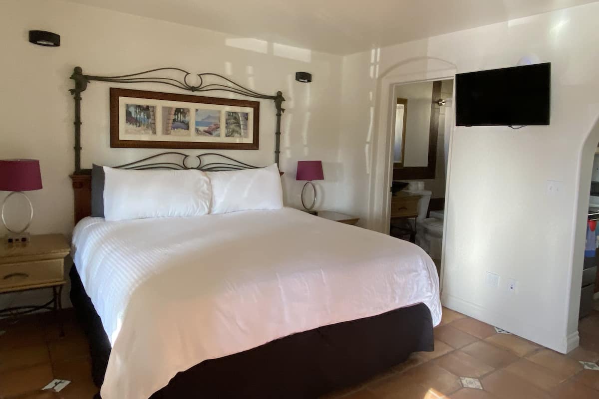 Ein Hotelzimmer mit einem Bett, zwei Nachttischlampen, einem gerahmten Bild über dem Bett, einem flachbildfernseher an der Wand und einem durch einen Bogen sichtbaren Badezimmer.