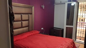 Ein Schlafzimmer mit Bett, Nachttisch, Schminktisch, einem Fenster mit Metallgitter und einem Deckenventilator.