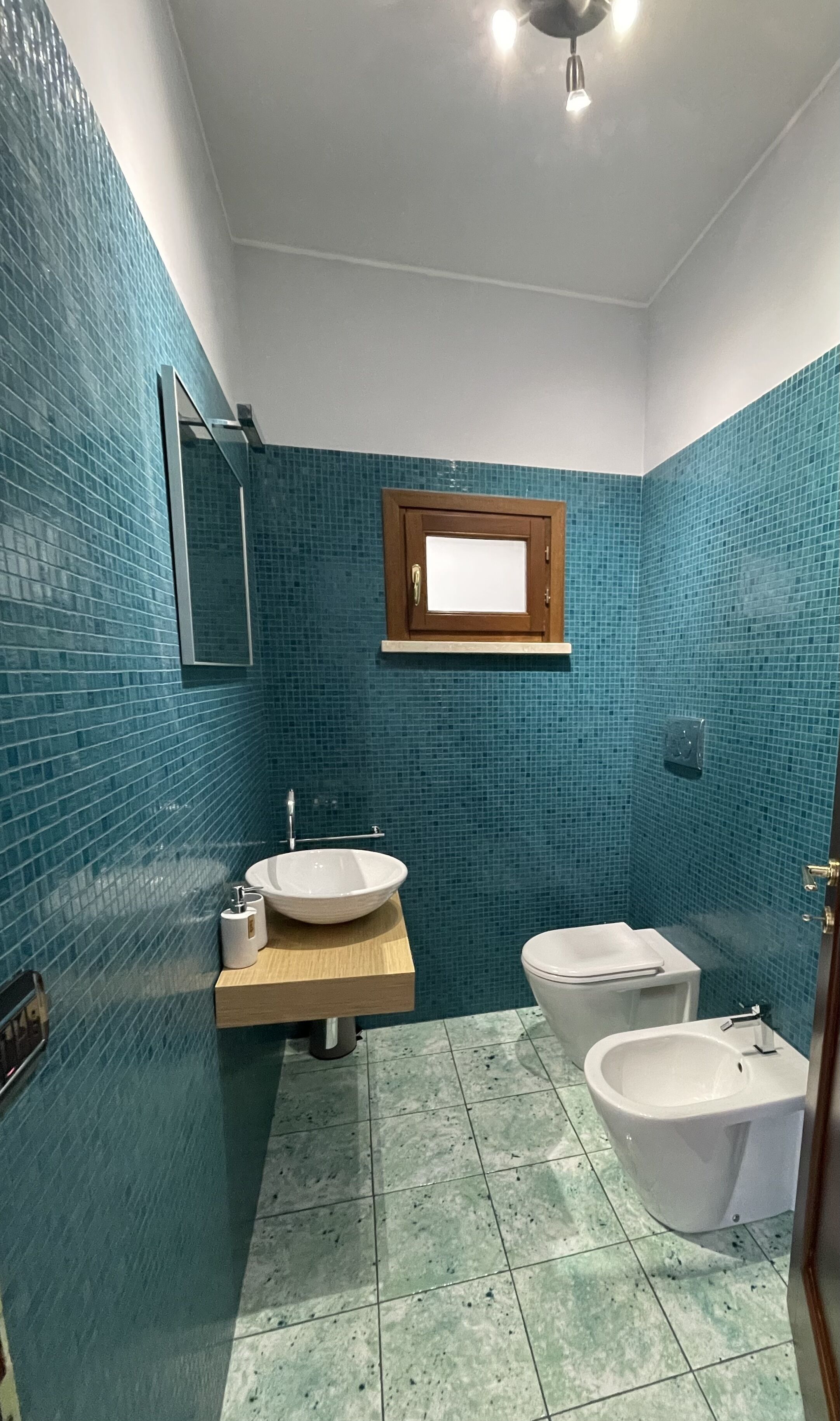 A bathroom with a sink, toilet, and bidet, tiled in blue, with a wooden vanity and a mirror above the sink.