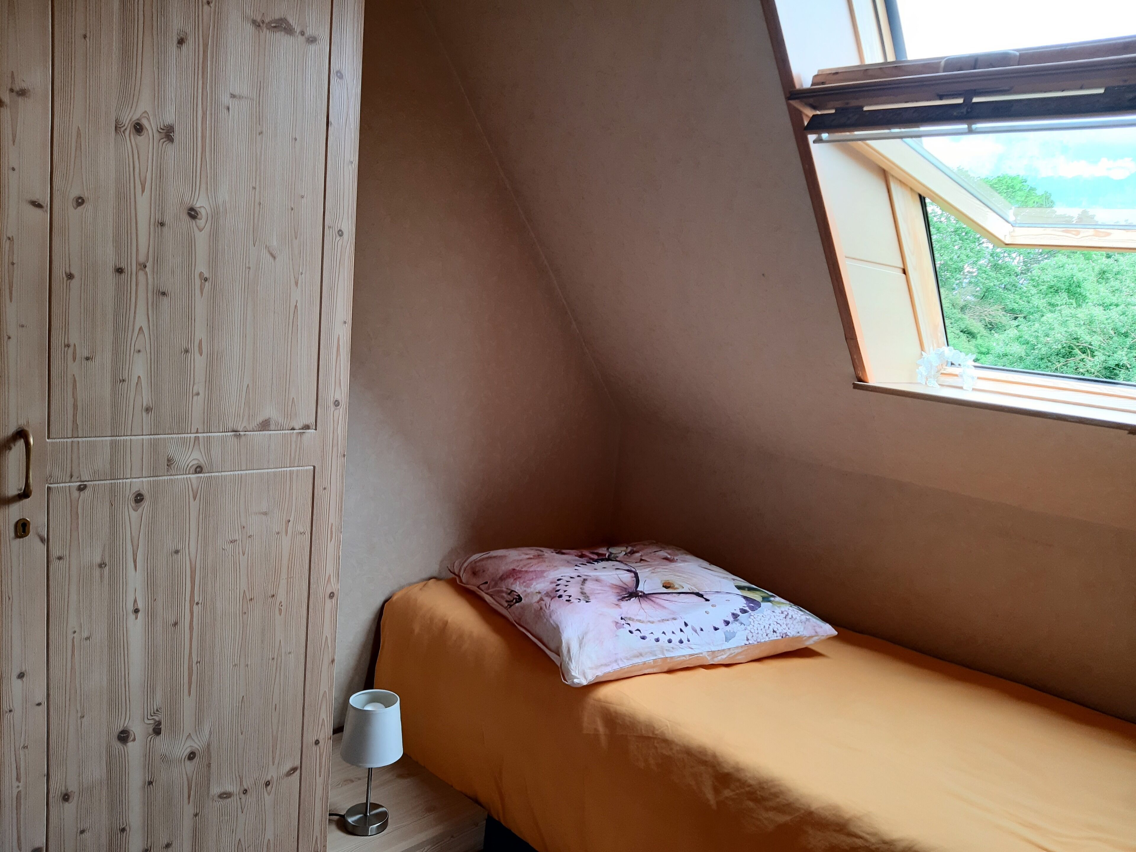 Un dormitorio con una cama individual, un armario de madera, una ventana con vistas a la vegetación y una mesita de noche con una lámpara.