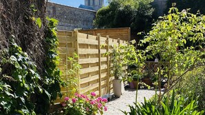 A cobblestone pathway leading to a wooden gate, surrounded by lush greenery and flowering plants.