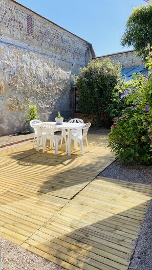 Een houten terras met een witte tafel en stoelen, omgeven door groen en een stenen muur.
