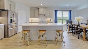 A modern kitchen with a central island, bar stools, and a dining area with chairs and a table. - New home, Argyle TX, 4 Bd rooms, 5 bds, 3 Bathrooms, Office, Gameroom, Sleeps 11 (Argyle)