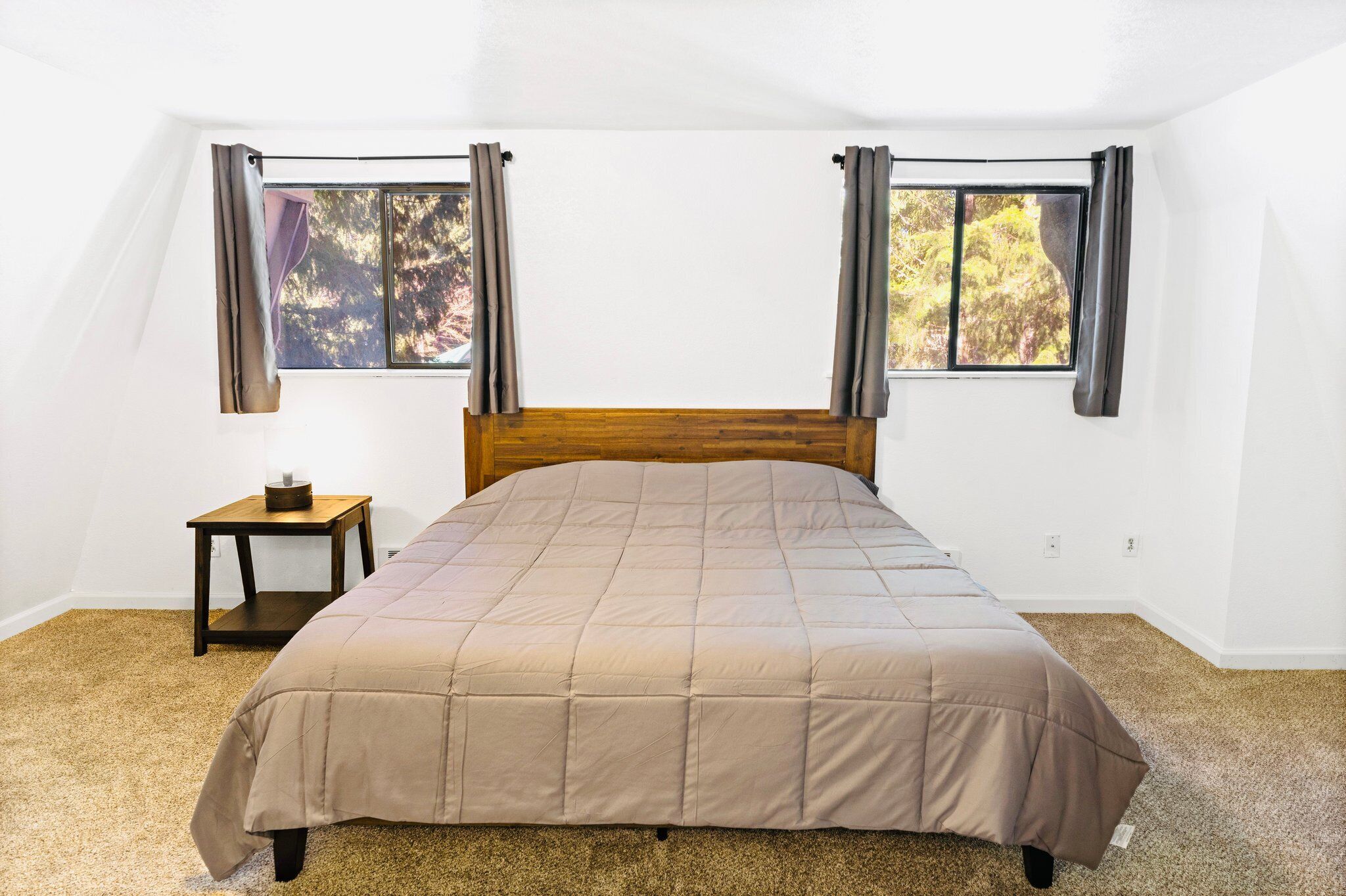 A bedroom with a wooden headboard, a beige bedspread, a small wooden nightstand, and two windows with curtains.