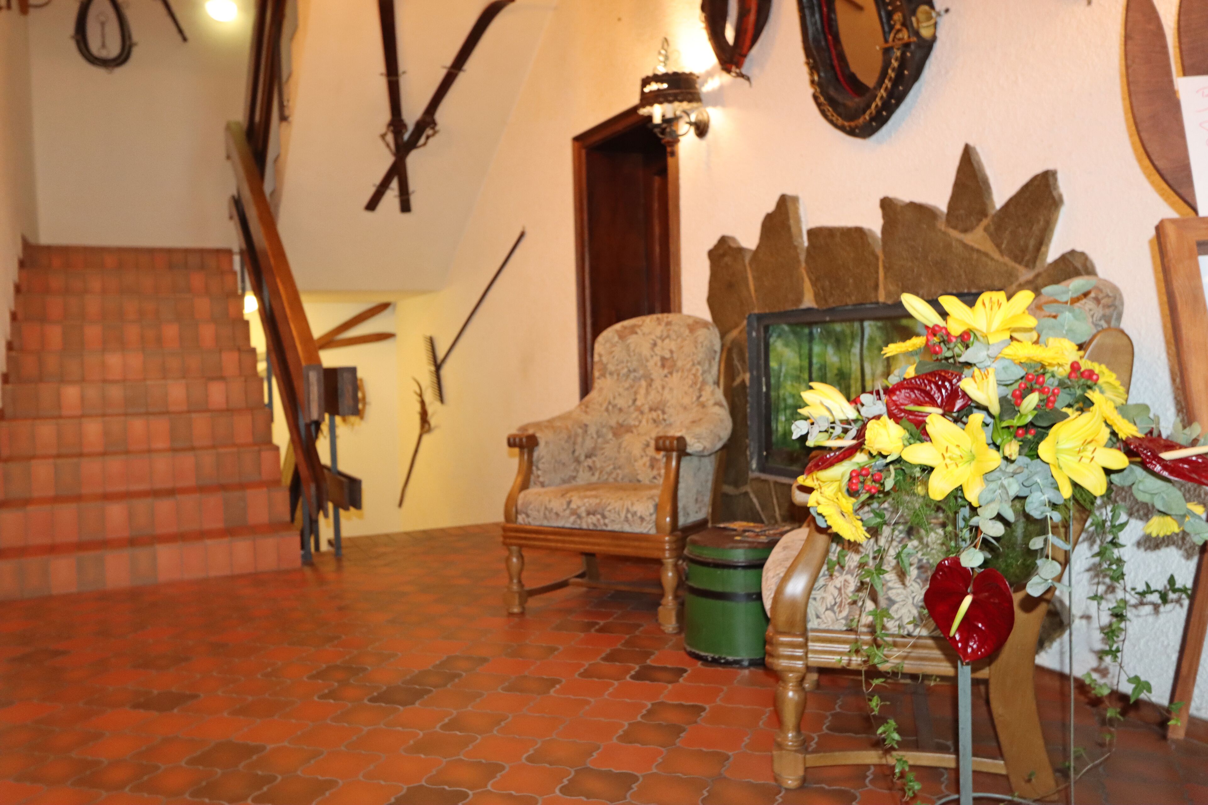 A room with a wooden chair, a green trunk, and a floral arrangement on a stand.