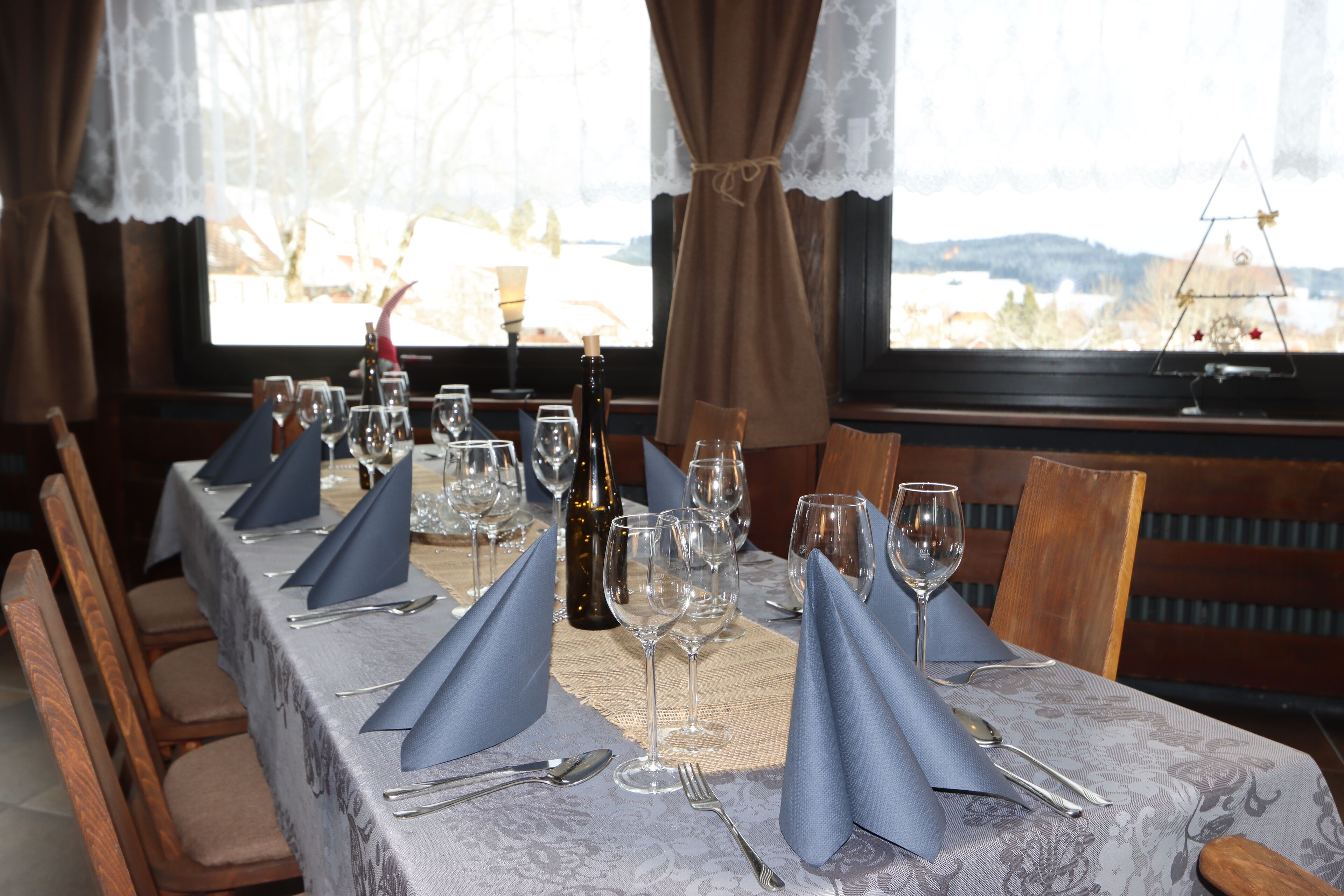 Una mesa de comedor dispuesta con servilletas azules, copas de vino y cubiertos, con vista a un paisaje nevado a través de una ventana.