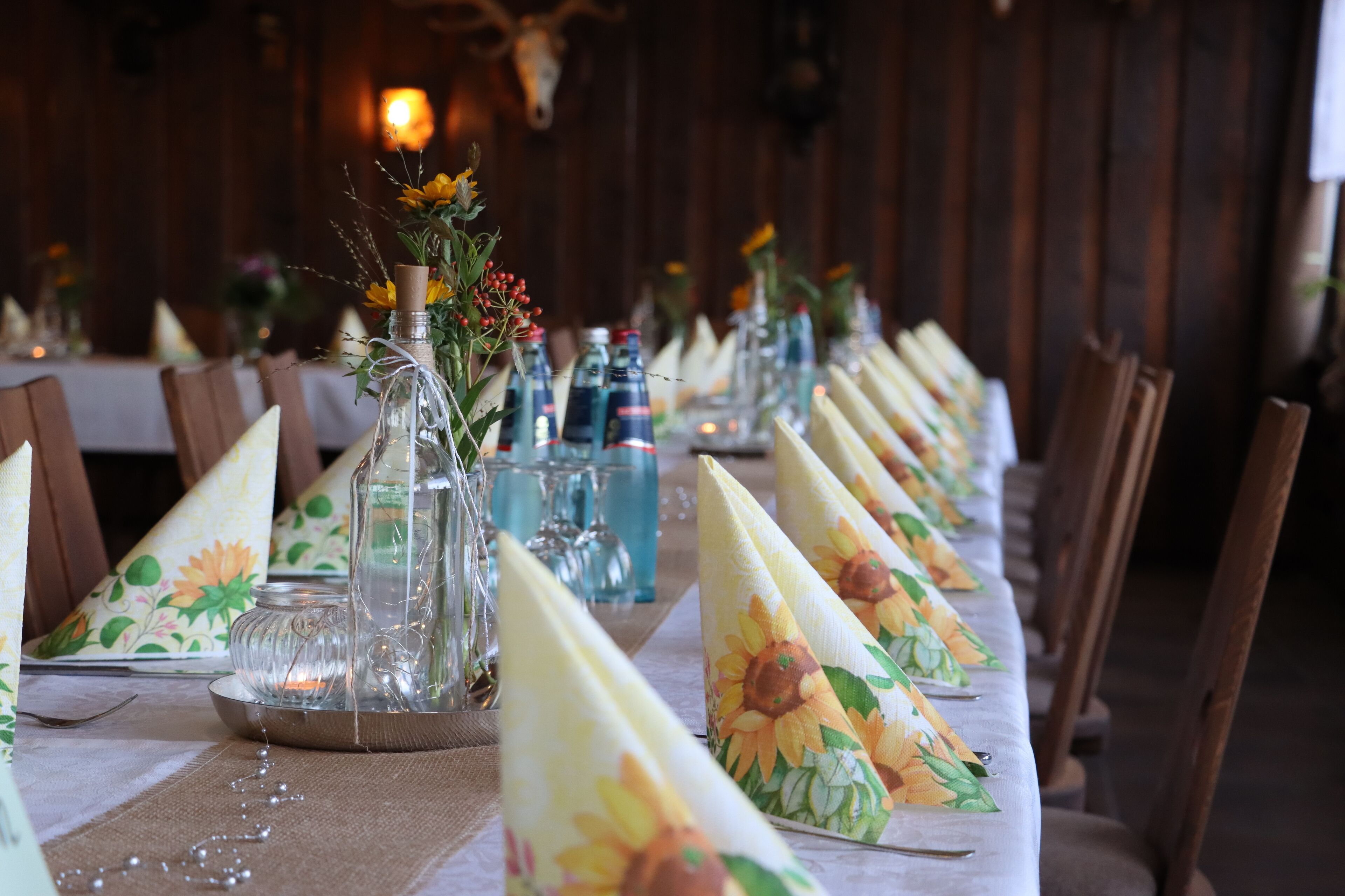 Una larga mesa de comedor decorada con servilletas florales, jarrones de vidrio con flores y vasos de cristal.
