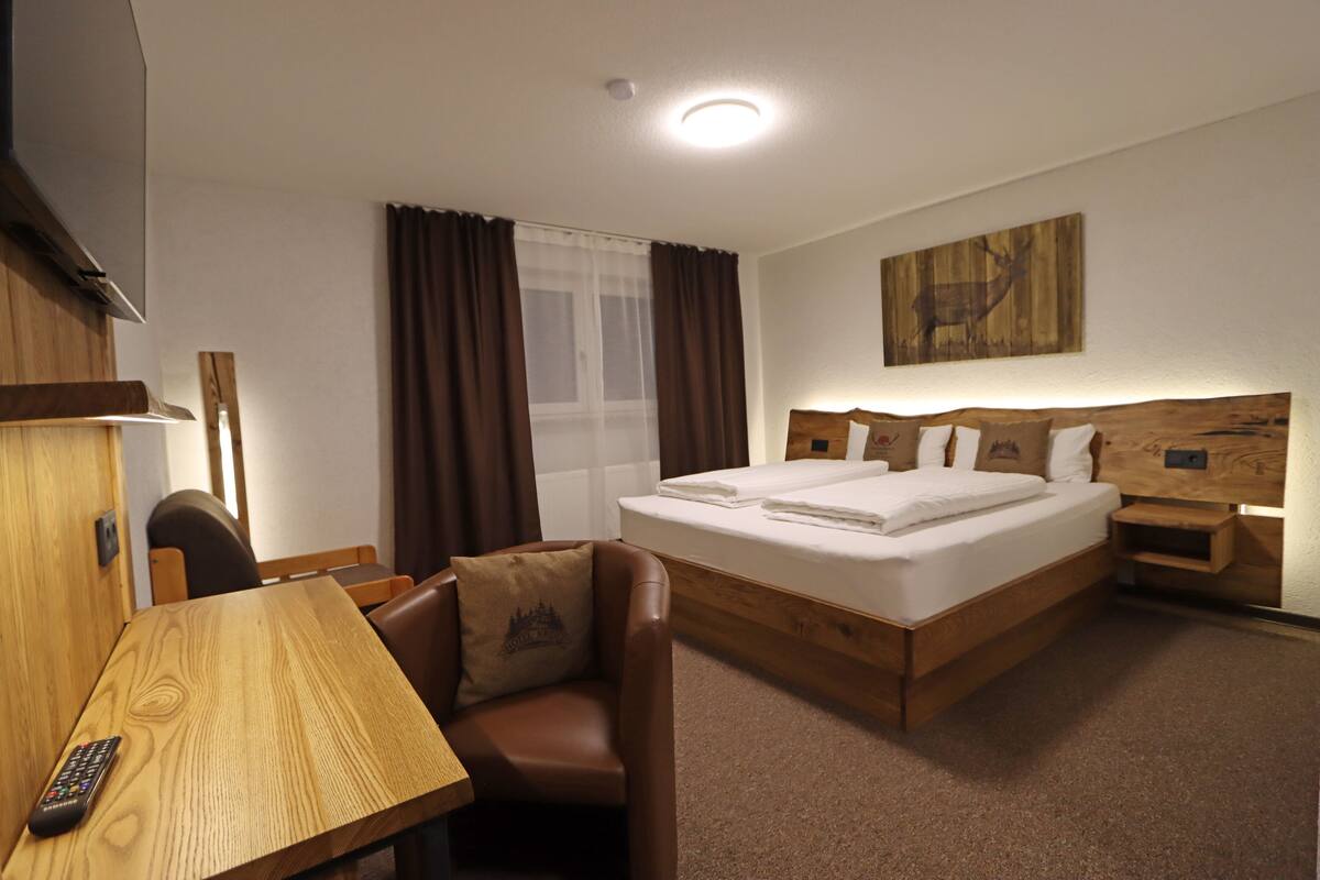 Une chambre d’hôtel avec un lit en bois, un bureau avec une télécommande, une chaise et une grande fenêtre avec des rideaux.