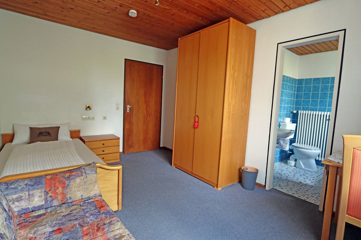 Une chambre à coucher avec un lit, une armoire, une petite table et une salle de bain visible à travers une porte ouverte.