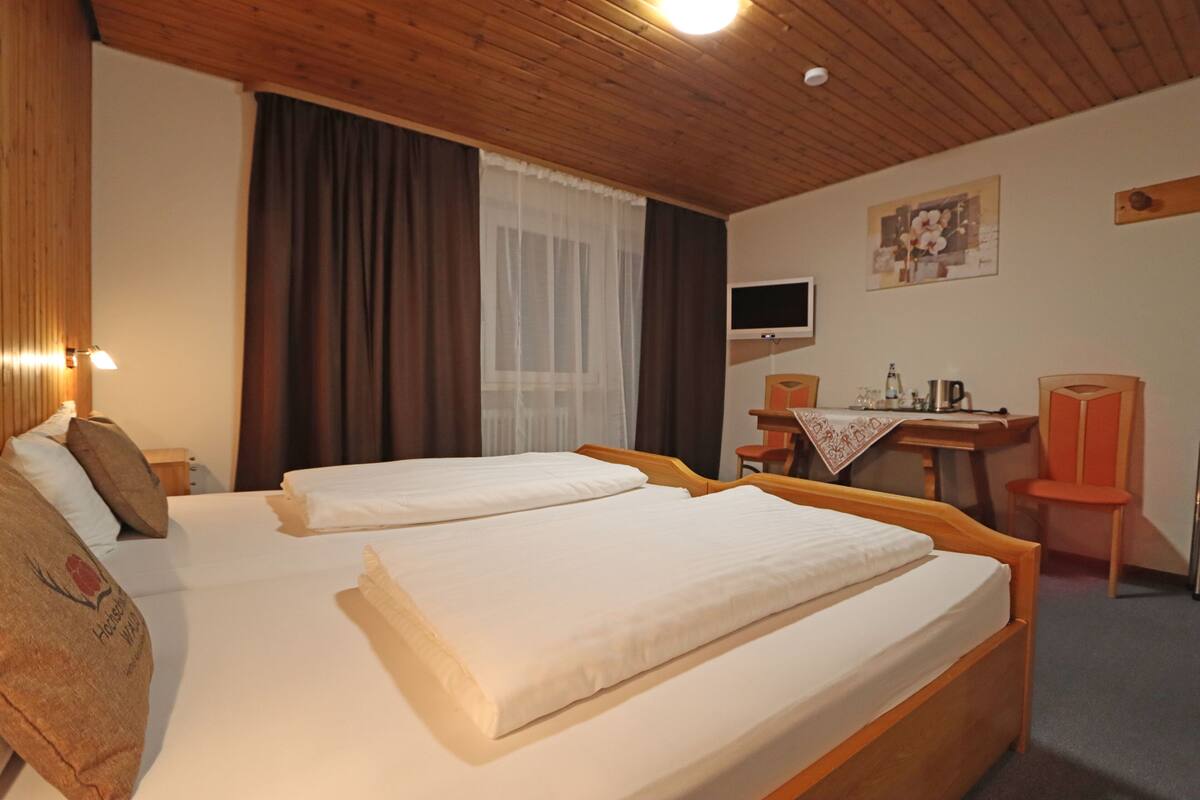Une chambre d’hôtel avec deux lits en bois, un bureau avec une chaise, une télévision et une fenêtre avec des rideaux.