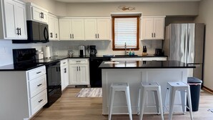 A modern kitchen with white cabinetry, a black countertop, a stainless steel refrigerator, and a window with blinds.