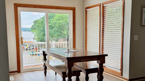 A room with a wooden table and chairs, a sliding glass door to a balcony, and a view of a body of water and greenery.