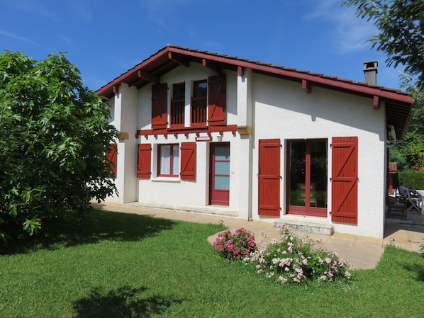 A two-story house with red shutters, a garden with pink flowers, and a clear blue sky.