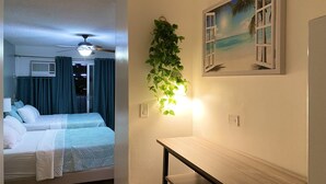Quarto de hotel com cama, escrivaninha, ventilador de teto, ar condicionado fixado na parede e janela com cortinas.