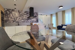 Un salon moderne avec une table basse en verre, un canapé et un coin cuisine doté de meubles blancs et d’un motif floral sur le mur.