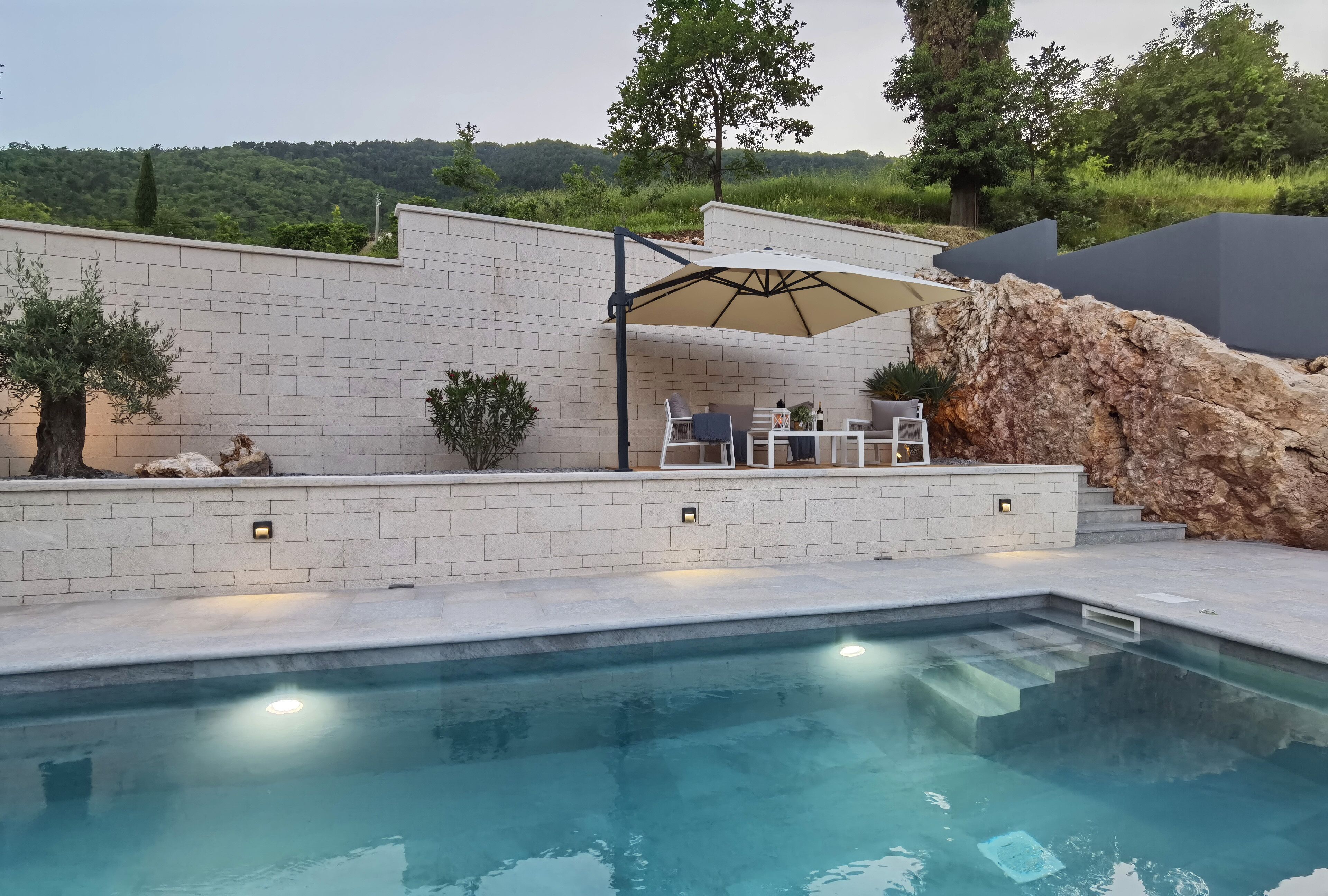 A swimming pool with a patio area, outdoor furniture, and a view of a hillside.