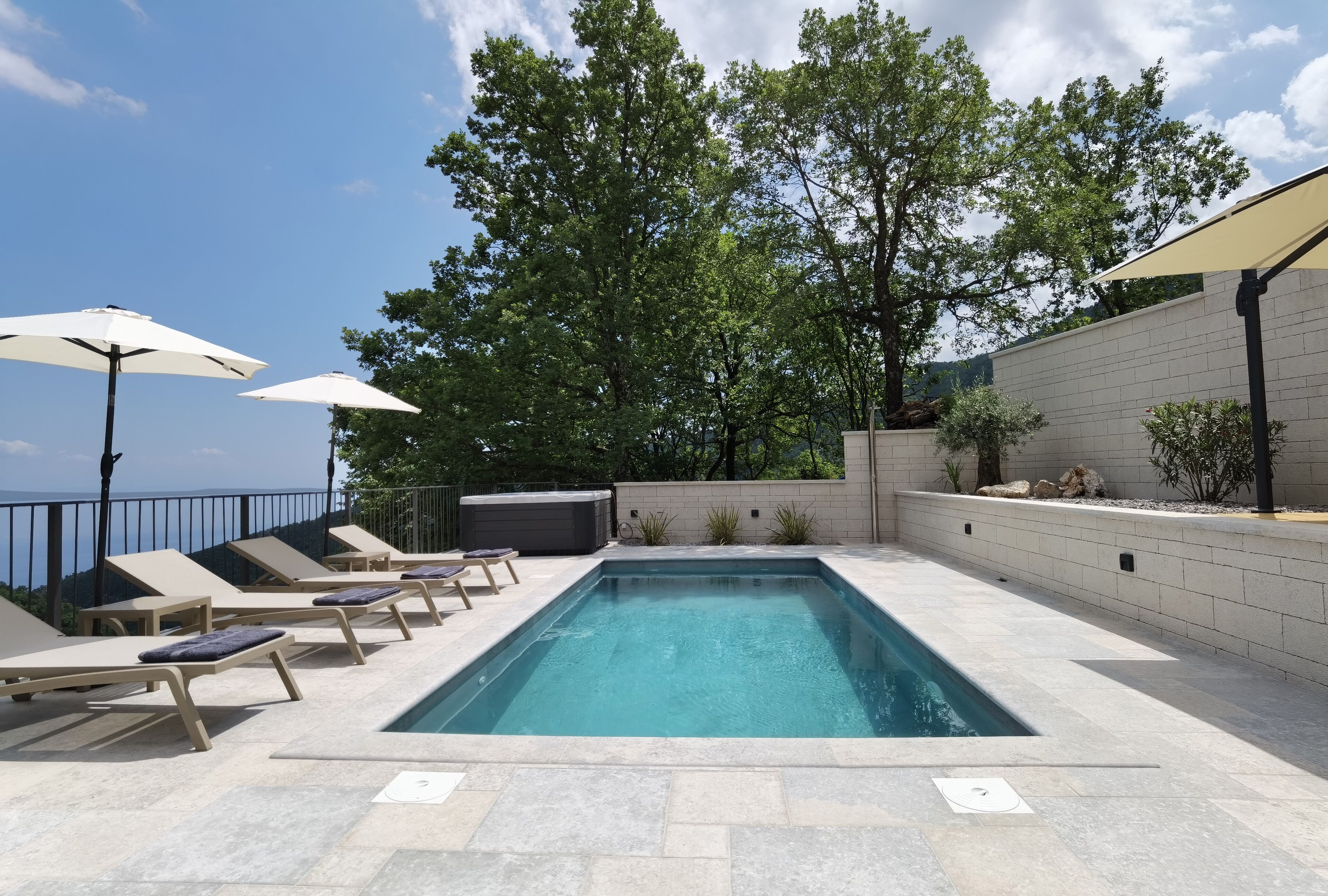 A rectangular swimming pool with lounge chairs and umbrellas, surrounded by a stone wall and trees.