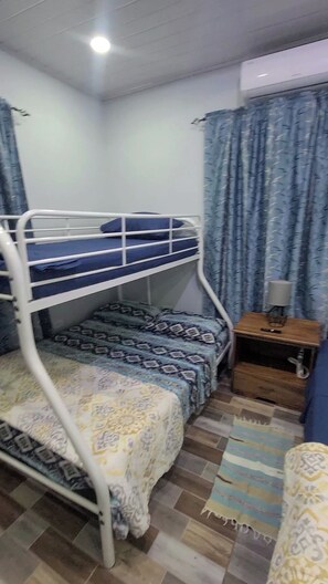 Een slaapkamer met een stapelbed, een bed met een patroon, een klein houten nachtkastje met een lamp en een vloerkleed met een dessin.