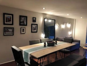 Un espace repas comprenant une longue table, des chaises et un canapé. Des tableaux encadrés ornent le mur, et un miroir est placé au-dessus de la table.