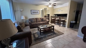 A living room with a sofa, coffee table, and lamp. There is a dining area with chairs and a table. The room has a ceiling fan and a kitchen area in the background. - 4-Person Condo only a short walk to beach. (Destin)
