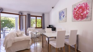 Un salon moderne avec un canapé, une table à manger et des chaises. De grandes fenêtres sont ornées de rideaux, il y a un téléviseur à écran plat et des œuvres d’art accrochées aux murs.