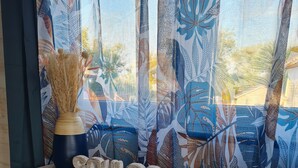 Ein Holztisch mit einer Vase und getrockneten Pflanzen, vor einem Fenster mit gemusterten Vorhängen.