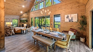 A spacious wooden cabin with a large dining table set for six, a cozy seating area, and a large window offering a view of the outdoors. - Playground|Views|Pets |Pool |Sauna |Massage Chair (Sevierville)