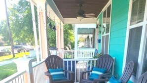Een overdekte veranda met houten vloer, een witte balustrade en drie Adirondack-stoelen met blauwe kussens.