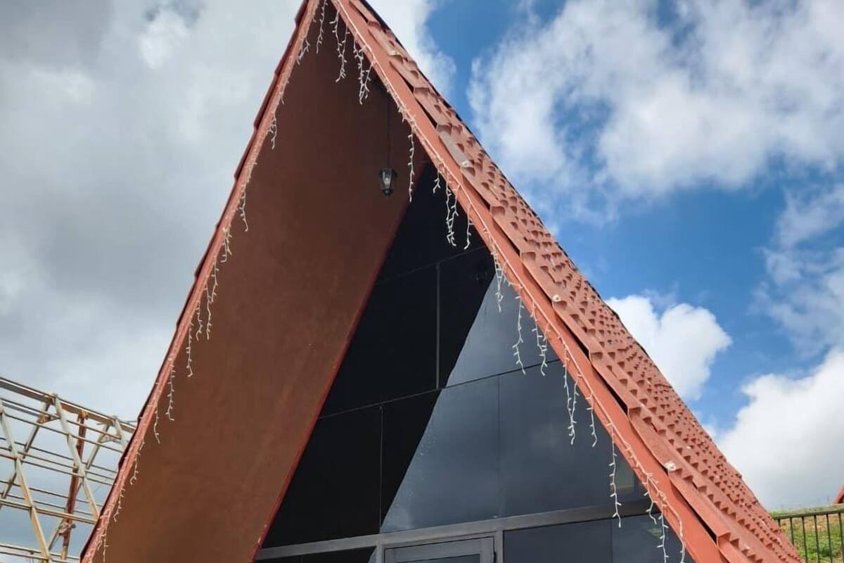 Una estructura moderna con techo triangular y pared de vidrio, un área de descanso con cojines y una terraza de madera.