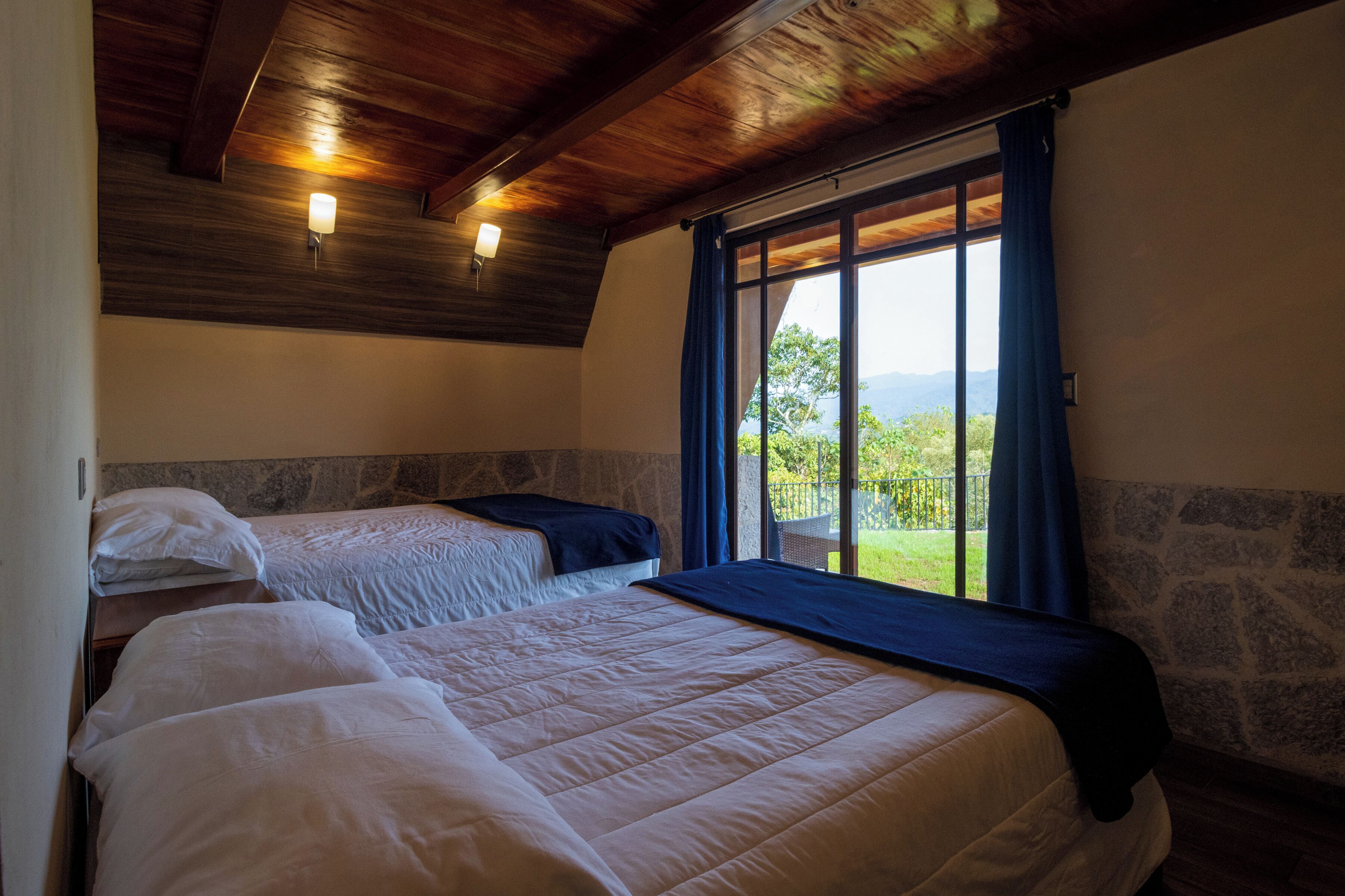 A room with two beds, a stone wall, wooden ceiling beams, and a large window with a view of greenery.